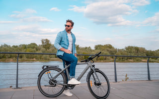A man wearing a denim shirt sitting on a grey ADO Air 30 Ultra city e-bike by a riverside promenade in the UK.