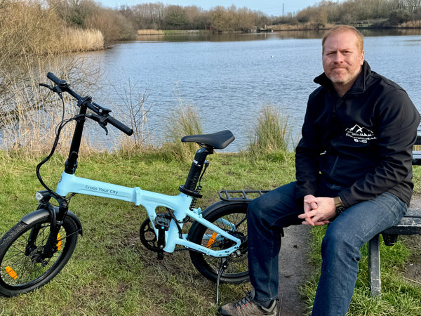 A man sitting on a wooden bench by a lake in the UK with an ice blue ADO Air 20 Pro folding electric bike parked beside him.