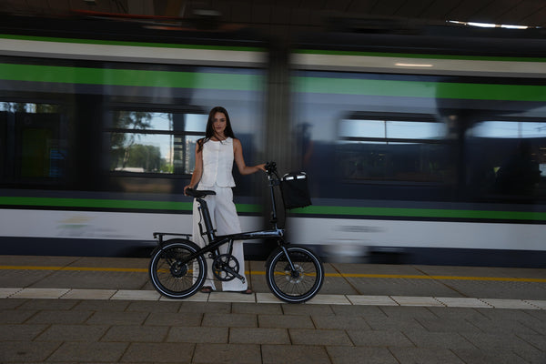 A woman standing on a railway station platform next to a black ADO Air Carbon Pro folding carbon electric bike with a front basket, as a train passes by.