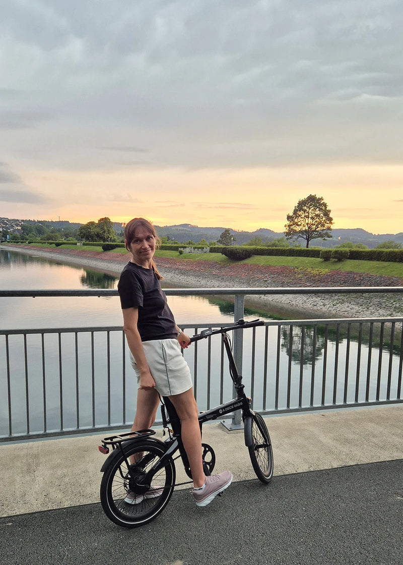 Woman standing with a folding ebike by a water railing at sunset.